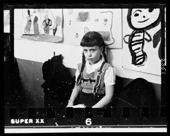 Young girl, half-length portrait, standing against wall displaying art work, in classroom in Chicago, Illinois